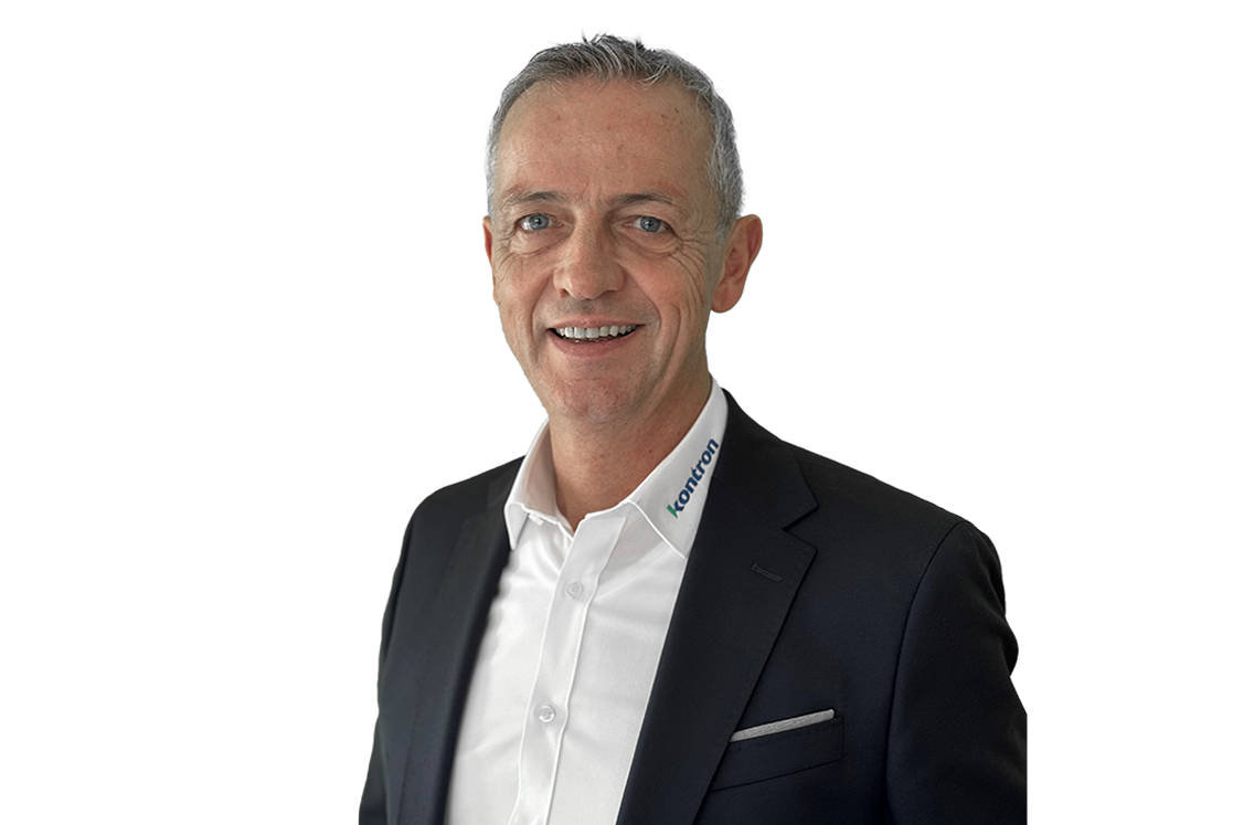 A smiling man in a black suit with a white shirt stands against a plain background. He has short, light-colored hair and is looking directly at the camera.