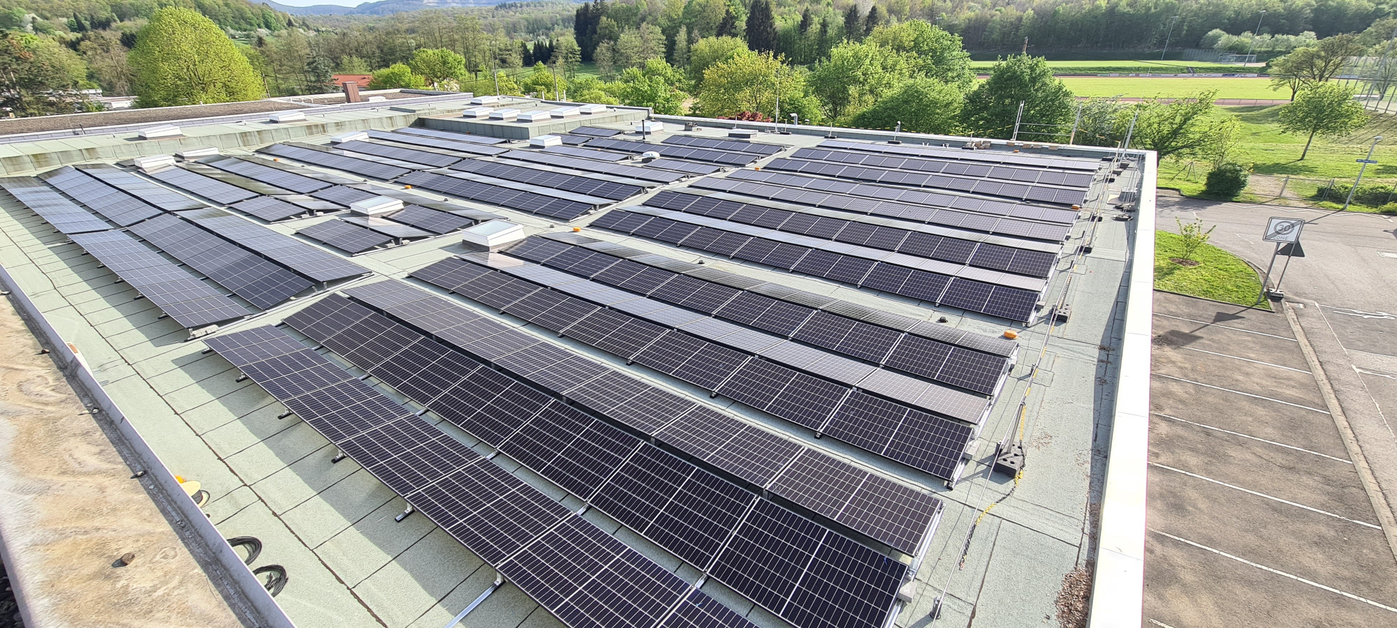 A large rooftop covered with numerous solar panels, surrounded by green trees and fields in the background.