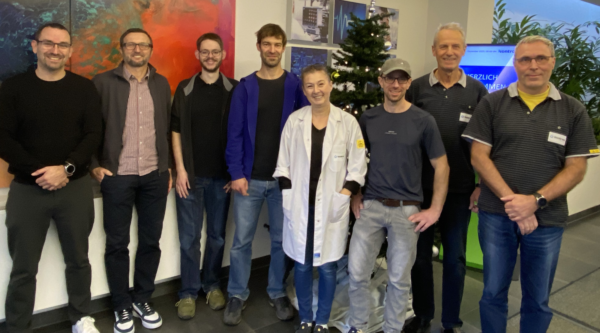 A group of eight individuals stands together in a brightly lit room decorated for the holidays, including a Christmas tree in the background. They are smiling and wearing a mix of casual and professional attire.