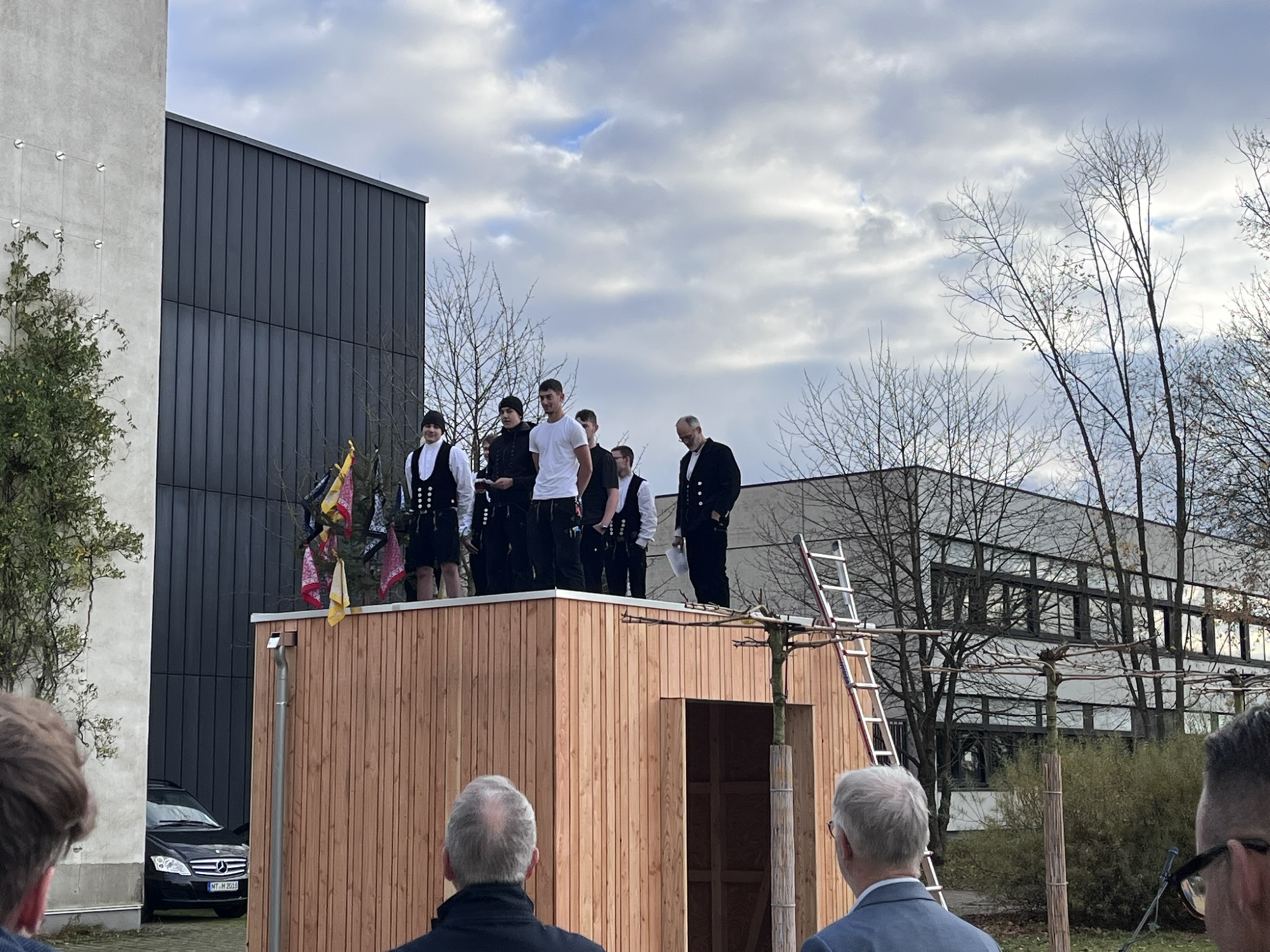 A group of people stands on top of a wooden structure, wearing traditional clothing. They are surrounded by trees and modern buildings. Some onlookers are visible in the foreground, observing the scene.