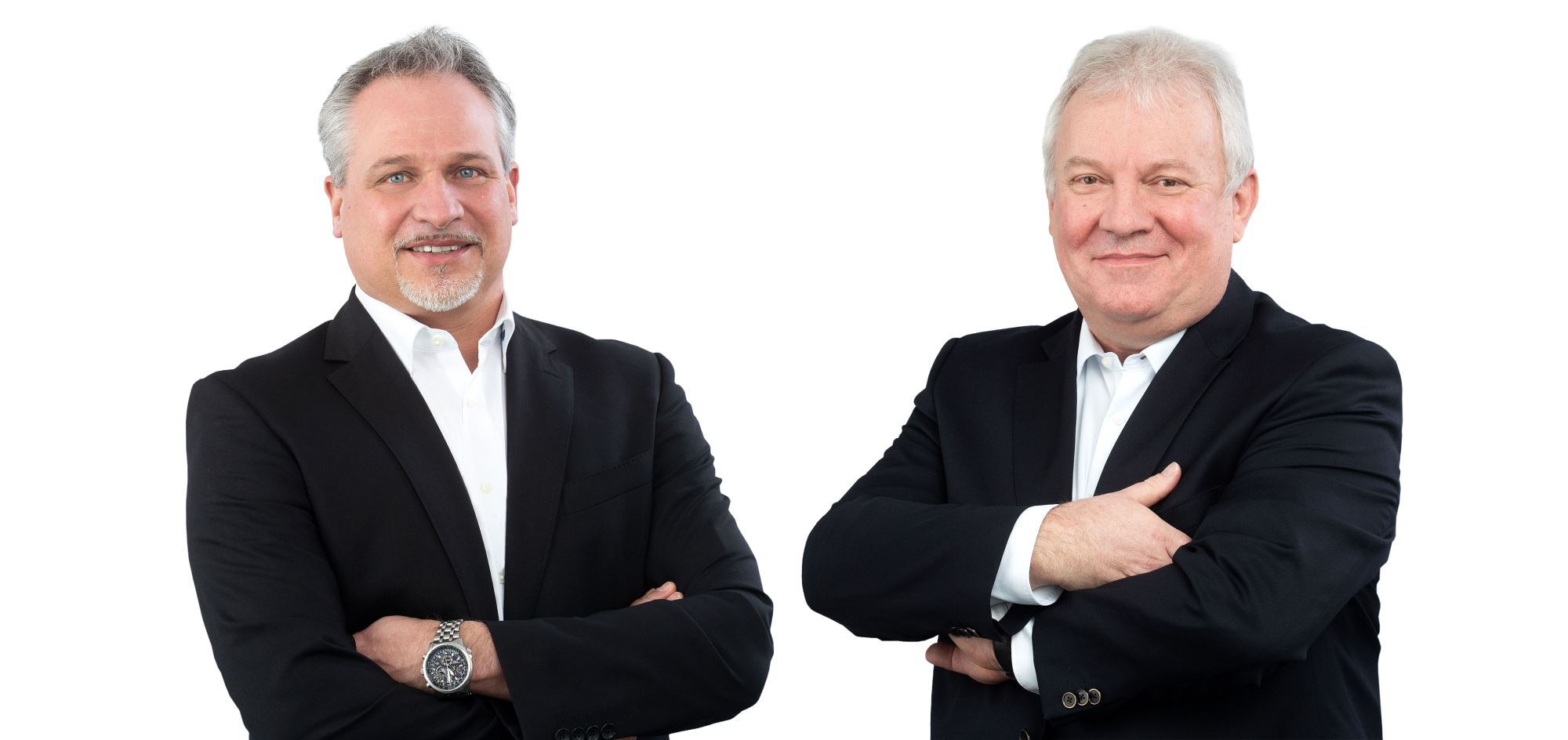 Two men in black blazers with white shirts, standing side by side. One man has gray hair and a beard, while the other has white hair and a more serious expression. They are both smiling slightly and have their arms crossed.