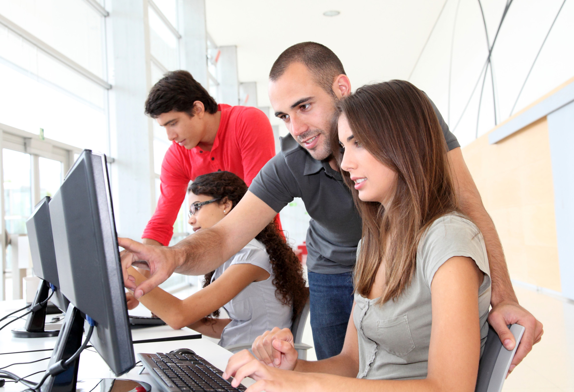 A group of four young adults is collaborating in a modern workspace. One man is pointing at a computer screen while assisting a woman who is typing. Two other women are focused on their own computers in the background. The environment features large windows and a bright, open design.