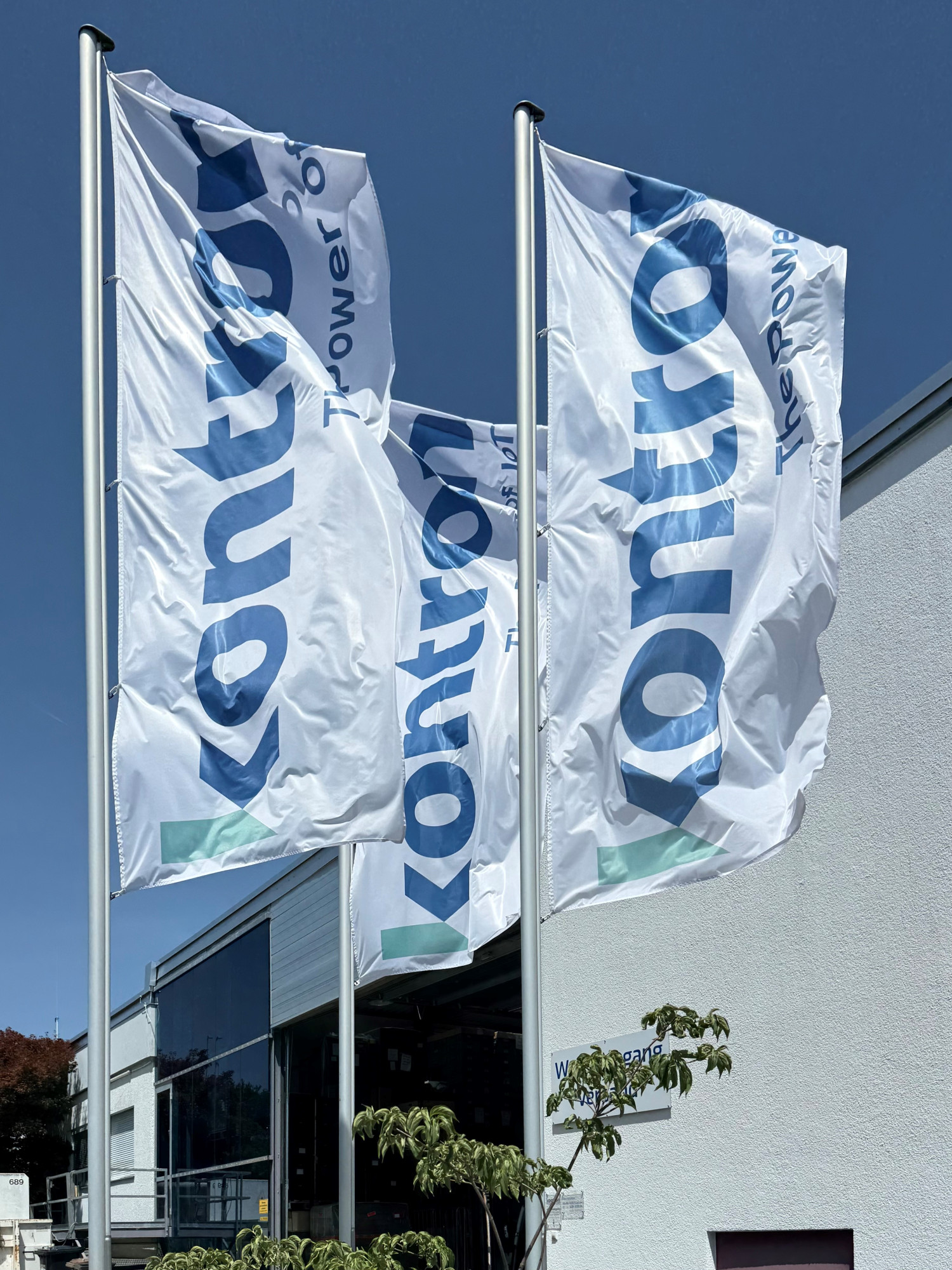 Three flags fluttering in the wind, displaying the word "kontrol" prominently. They are attached to metal poles in front of a modern building with a clear blue sky in the background.