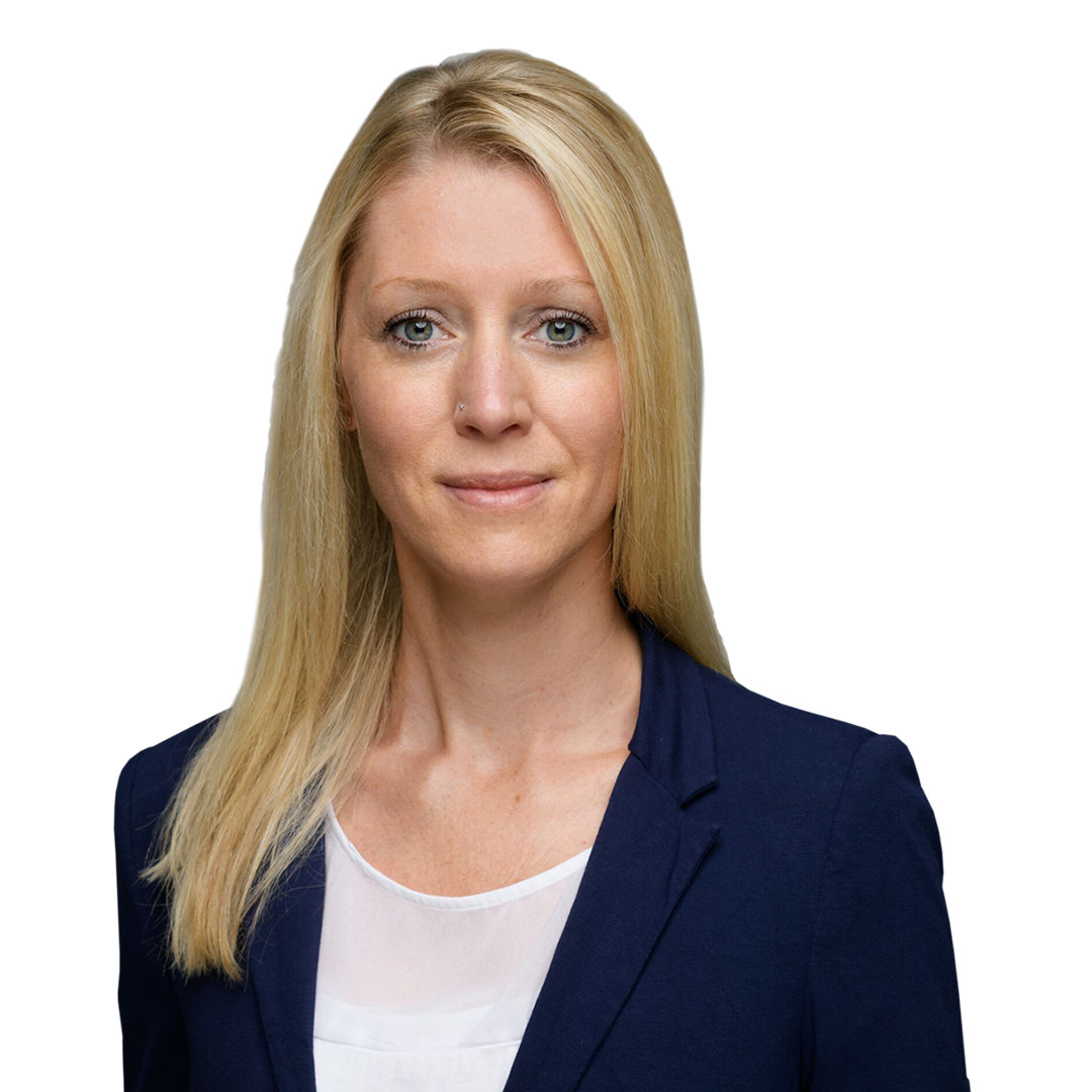 A woman with long blonde hair, wearing a dark blazer over a white blouse, stands against a plain white background. She has a neutral expression and direct gaze.