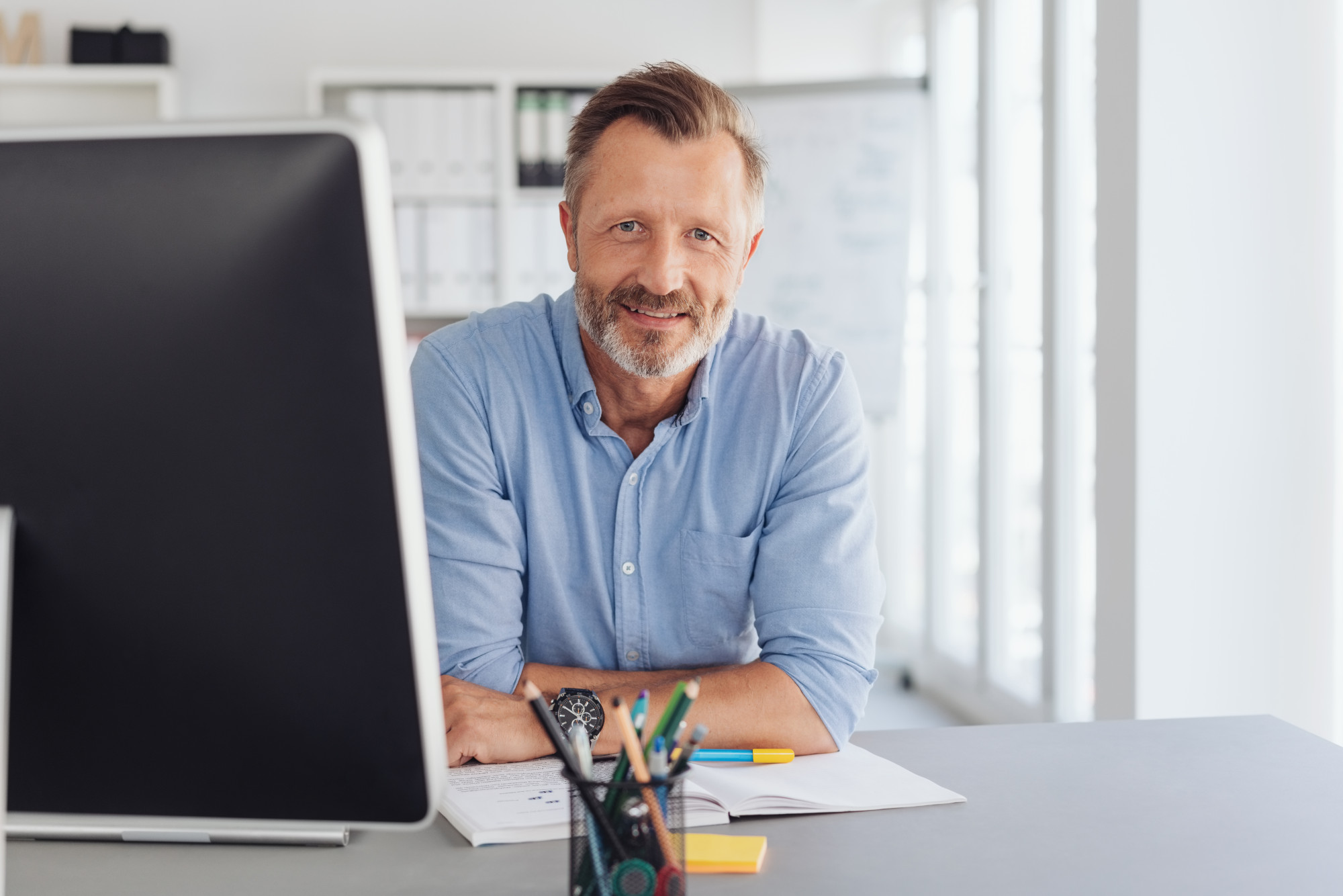 A man with a beard and friendly smile sits at a desk in an office, looking towards the camera. He is wearing a light blue button-up shirt and has a watch on his wrist. A computer monitor and stationery supplies are visible on the desk.