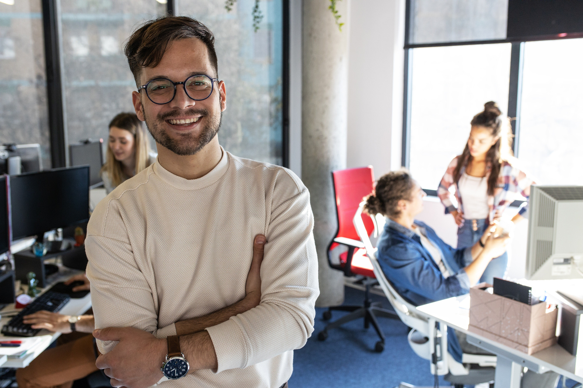 A smiling man with glasses stands confidently in an office setting, wearing a light-colored sweater. In the background, two women are engaged in conversation while another woman works at a computer. Large windows provide natural light and a view of the outside.