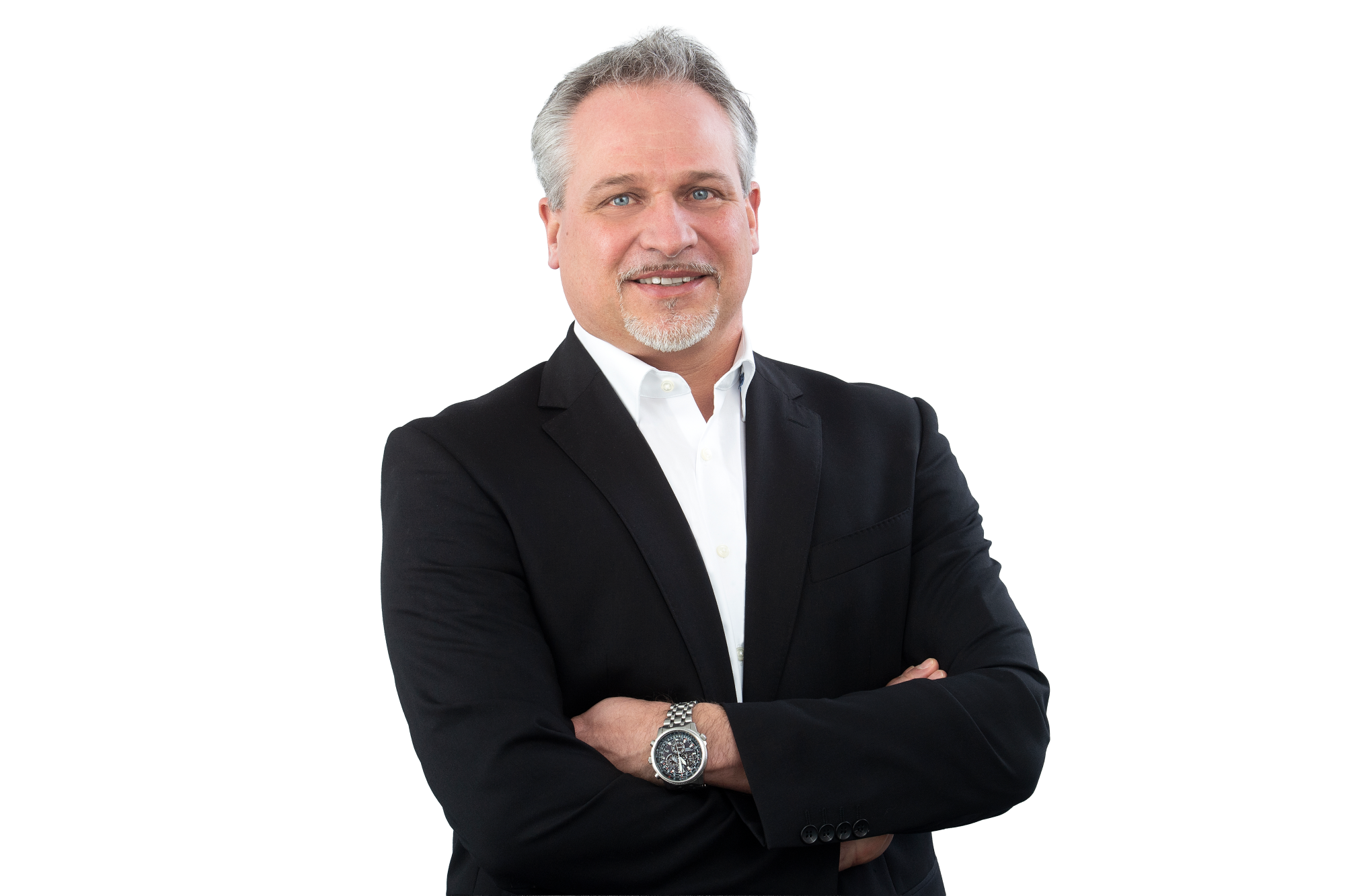 A confident man in a black suit and white shirt stands with arms crossed, smiling slightly against a white background. He has short, light-colored hair and is wearing a watch on his left wrist.