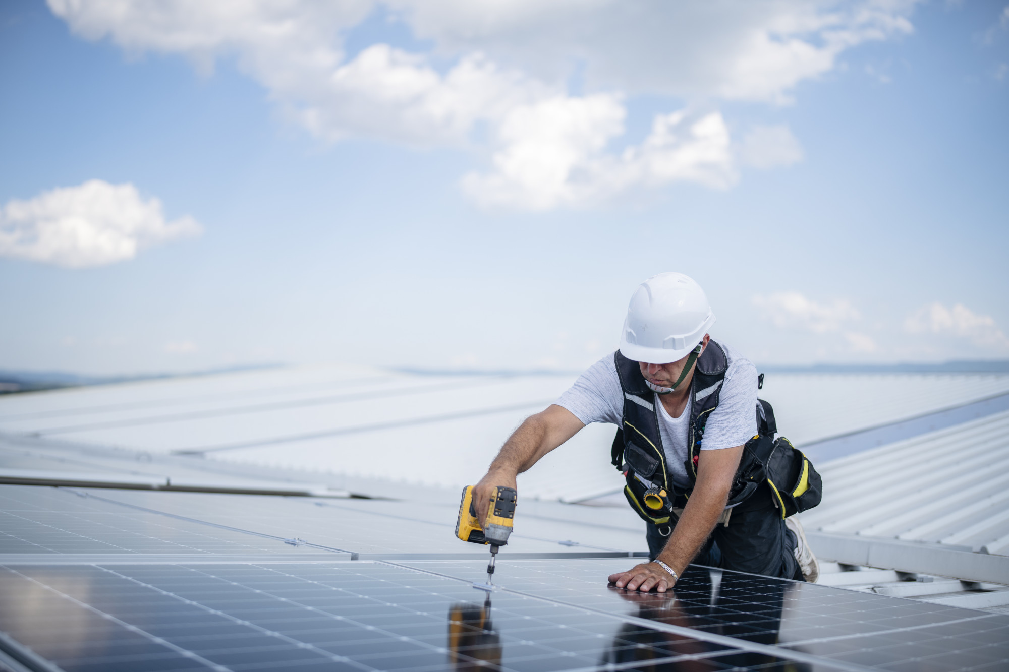 Ein Handwerker installiert eine Solaranlage auf dem Dach. Er trägt einen Helm und benutzt eine Bohrmaschine, um die Solarpaneele zu befestigen. Der Himmel ist blau mit vereinzelten Wolken.