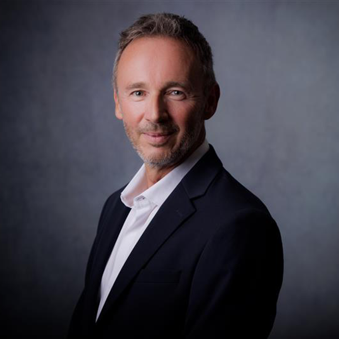 A man with short gray hair and a well-groomed beard is smiling confidently, wearing a dark suit jacket over a white shirt. The background is soft and slightly textured, enhancing the professional appearance.