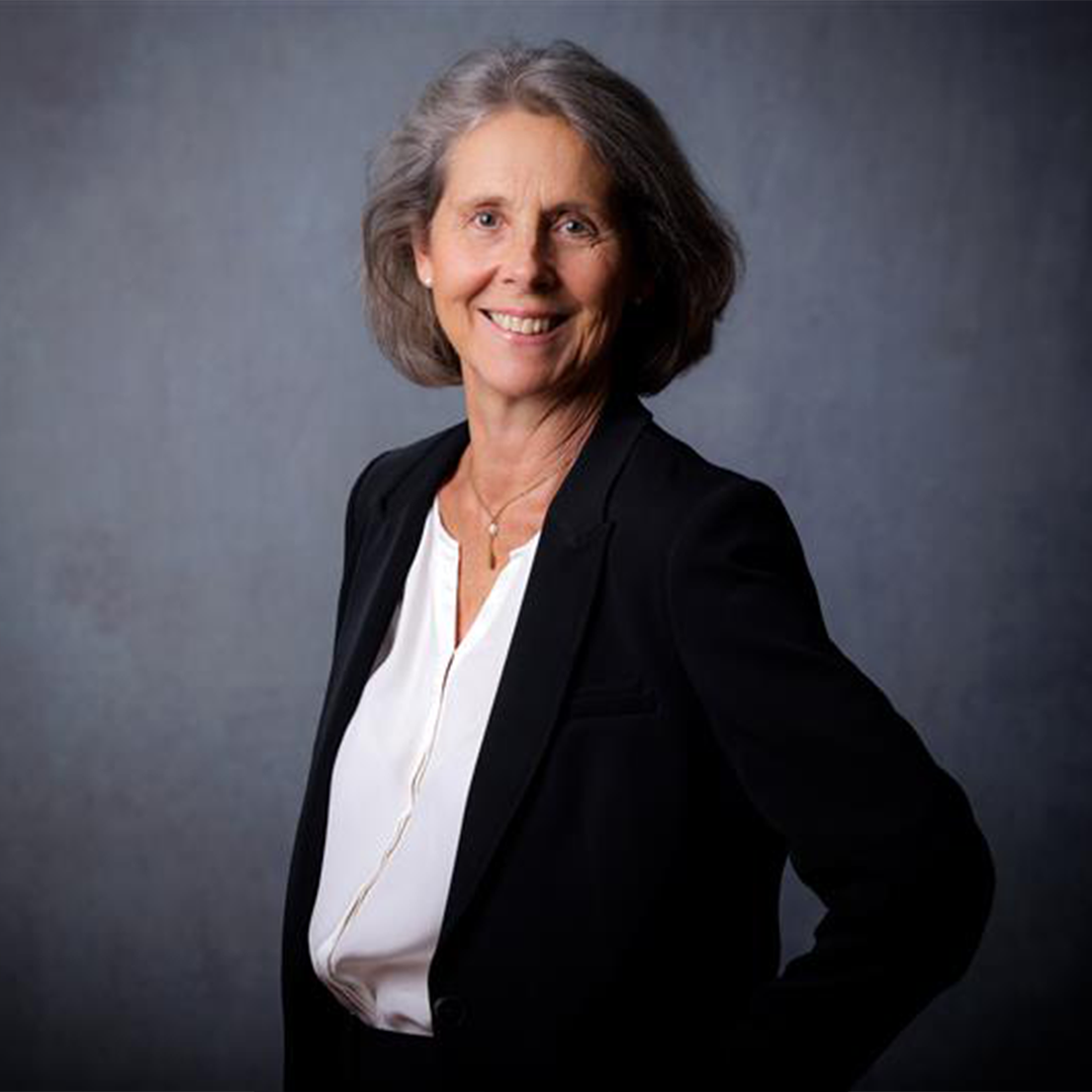 A woman with shoulder-length gray hair smiles while wearing a black blazer over a white blouse. She stands confidently with one hand on her hip against a textured gray background.