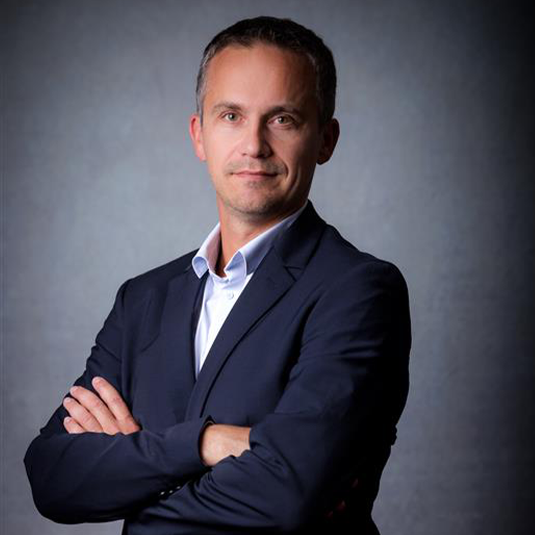A man in a dark suit stands confidently with his arms crossed. He has short, neatly combed hair and a serious expression, against a neutral gray background.