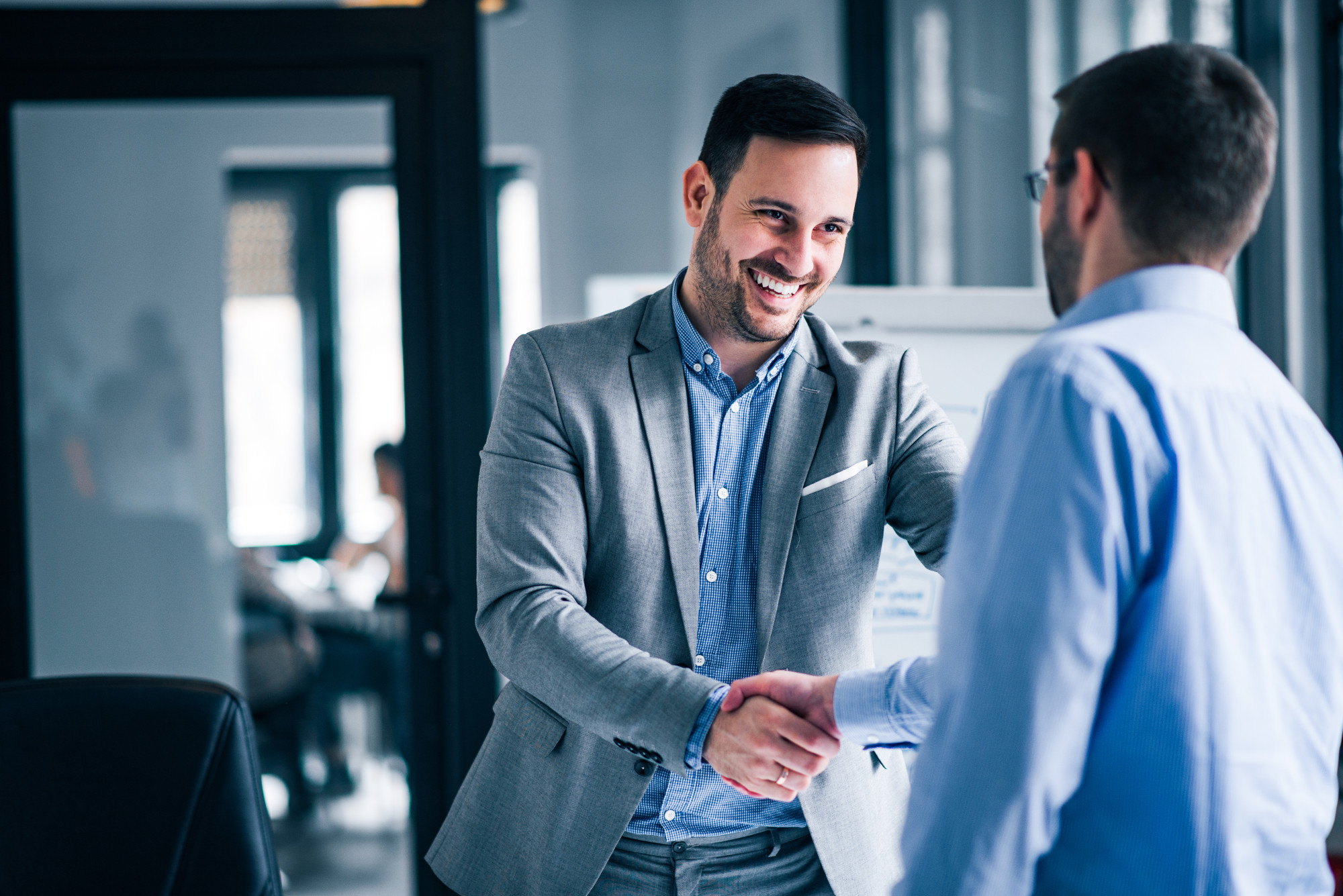 Two men in business attire shaking hands in a modern office setting. One man smiles while the other looks engaged. Background features glass walls and blurred office activity.