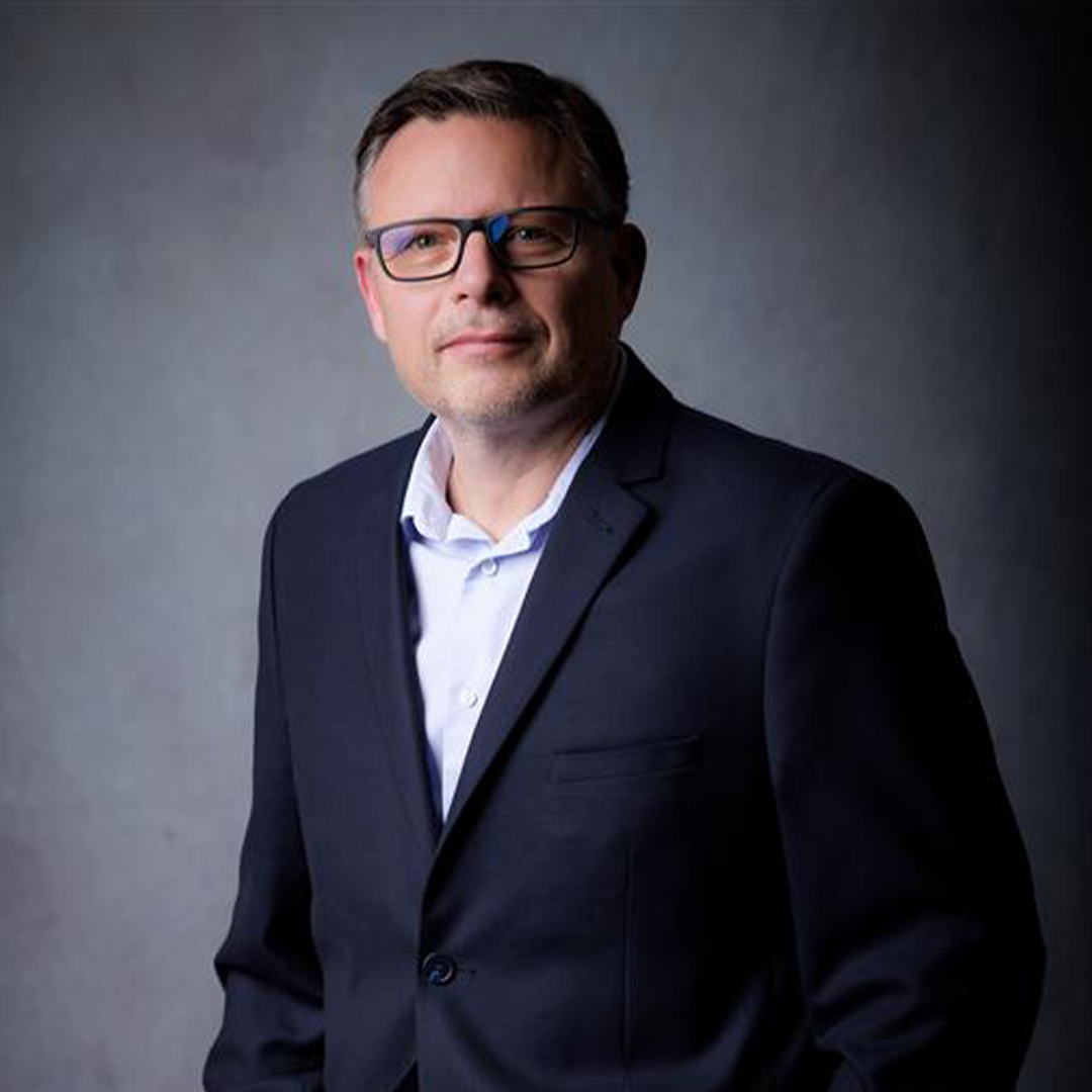 A man in a dark suit and light blue shirt stands confidently against a gray backdrop. He has short, dark hair and wears rectangular glasses.