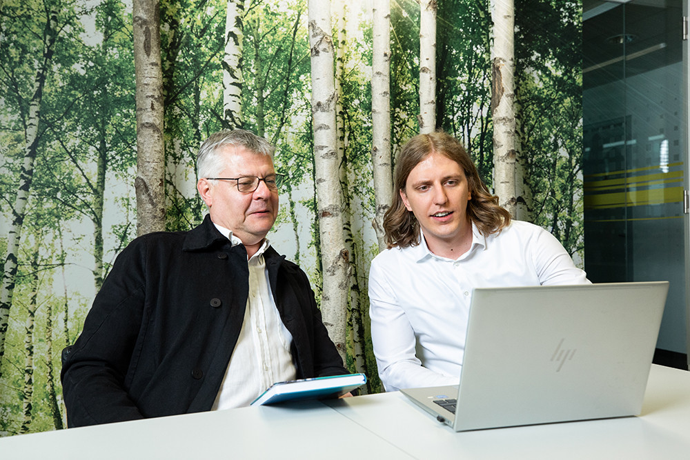 Two men are seated at a table, reviewing a laptop. One man has gray hair and glasses, while the other has long, light brown hair. They are in a bright room with a forest-themed wallpaper featuring birch trees.