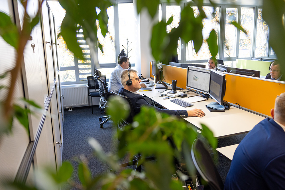 Several individuals are working in an office environment, wearing headsets and focused on computer screens. The workspace features modern cubicles with orange and white panels, and greenery is visible in the foreground.