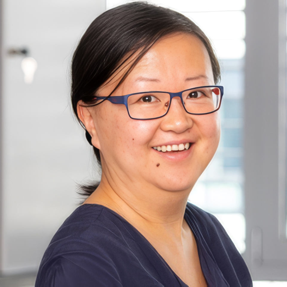Smiling woman with black hair and glasses, wearing a dark blue top, standing in a bright, modern indoor setting.