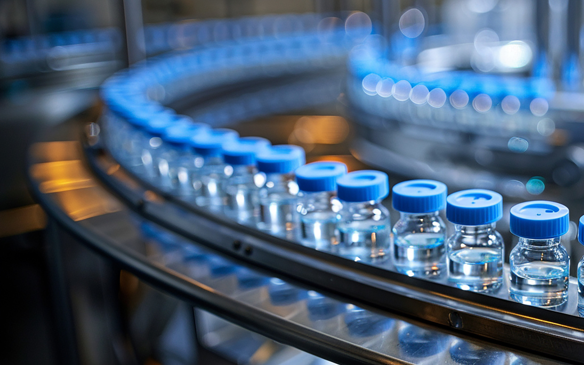 Row of small glass vials with blue caps, arranged on a conveyor belt in a laboratory or production facility. Blurry lights and equipment are visible in the background.