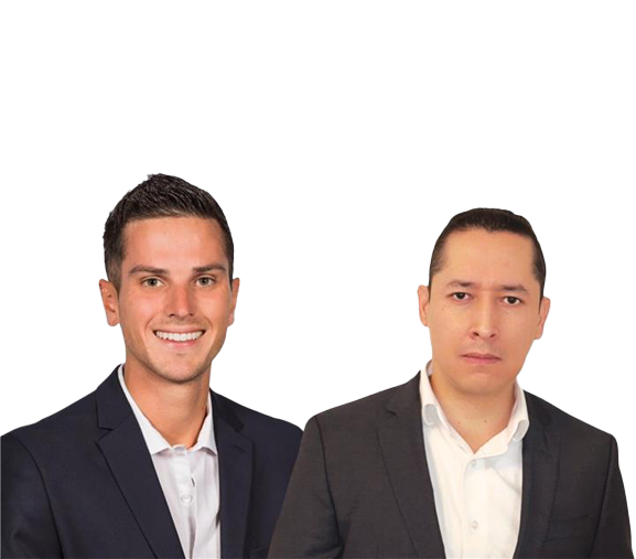 Two professionally dressed men stand side by side, smiling. The man on the left has short, dark hair and is wearing a navy suit with a white shirt. The man on the right has dark hair and a serious expression, dressed in a black suit with a white shirt. Both appear to be in a studio setting with a plain background.