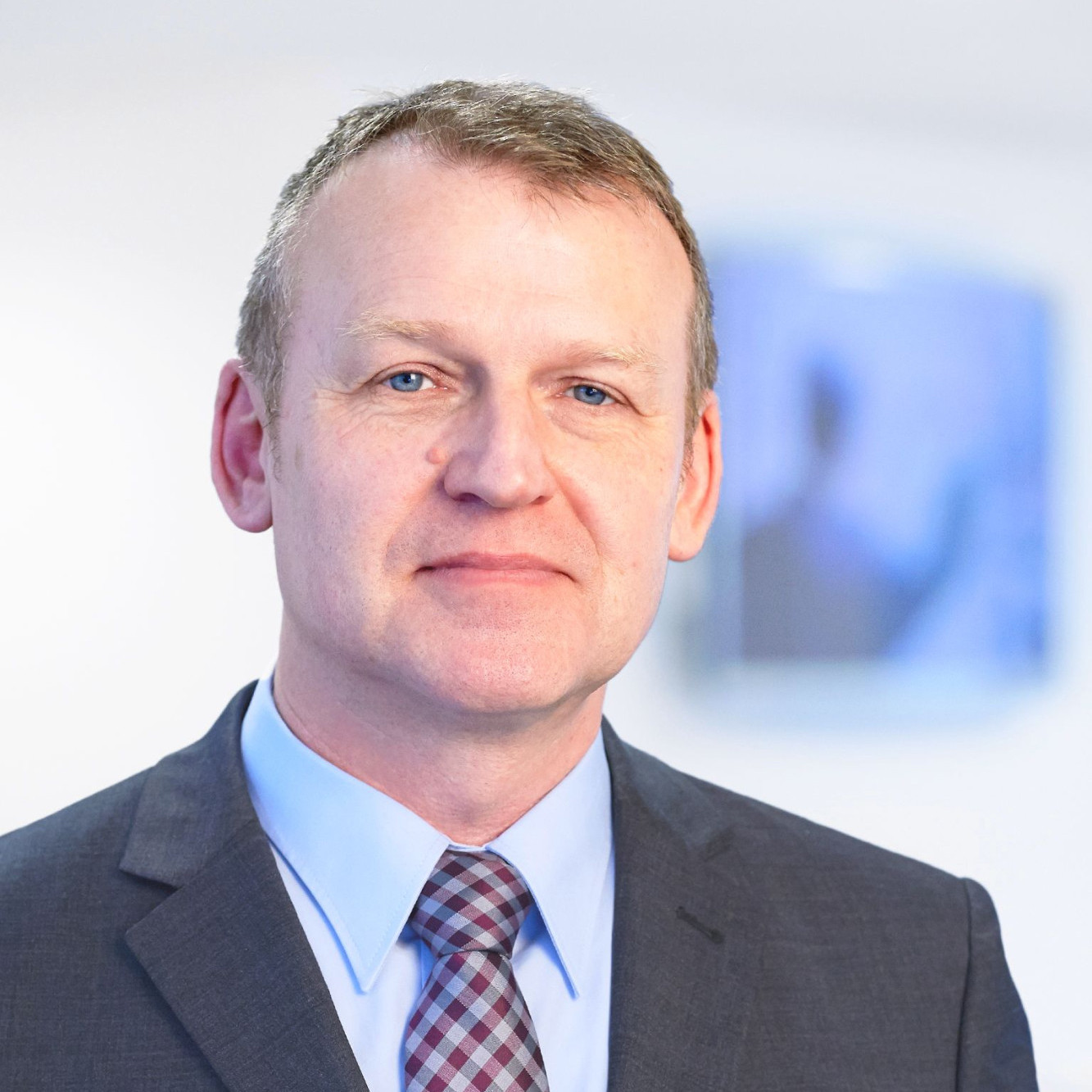 A man in a suit and tie smiles confidently, with short hair and blue eyes, standing in a professional setting. A blurred background suggests an office environment.
