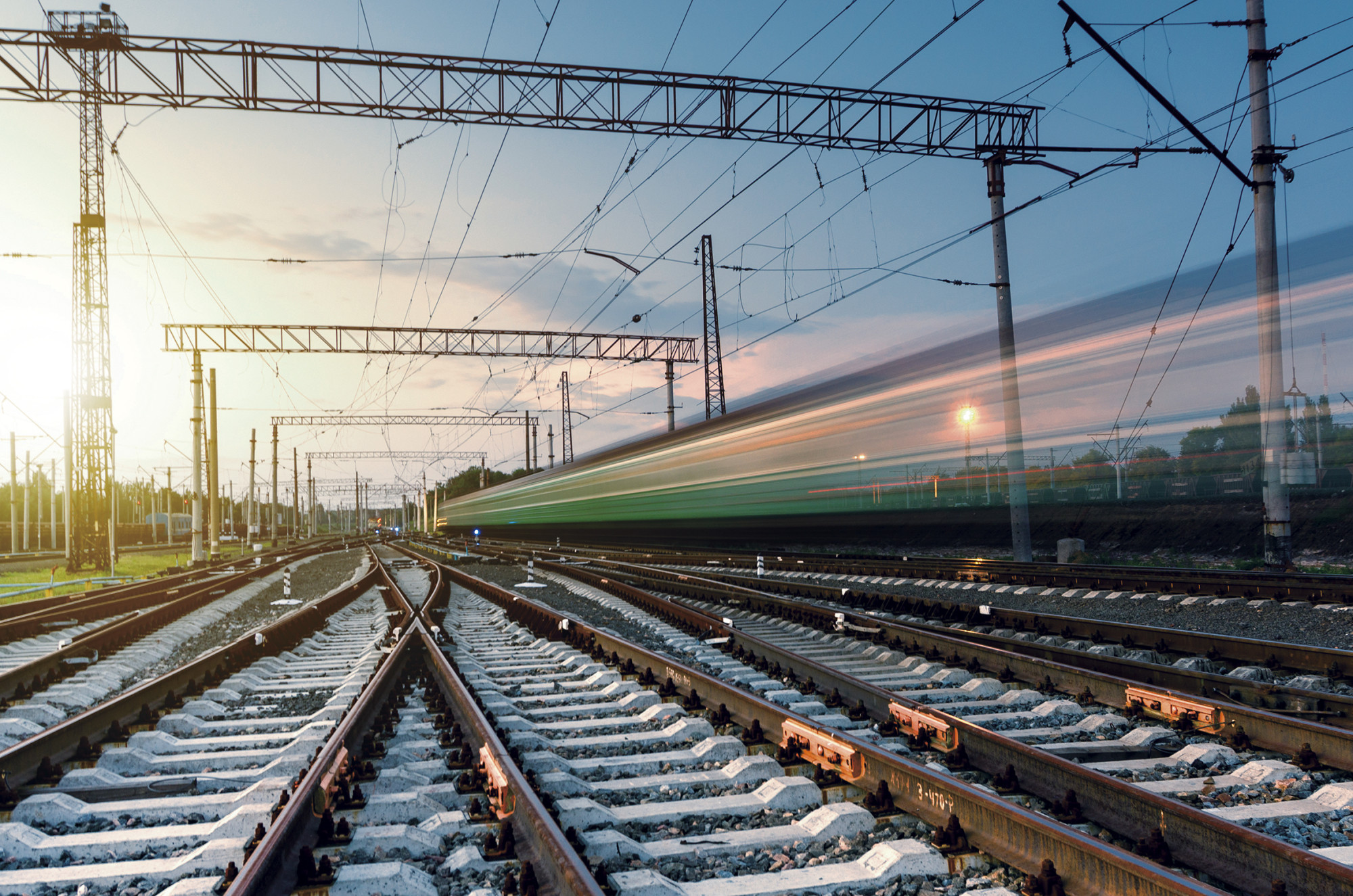 A sunset view of railway tracks with power lines, featuring a blurred train moving quickly through the scene.