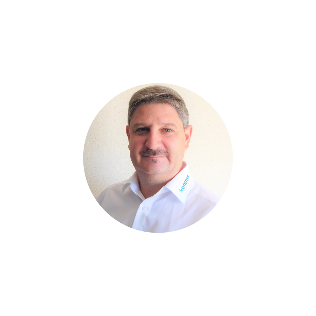 A man with short, light brown hair and a mustache is smiling while wearing a white collared shirt. The shirt has a branding label on the collar. The background is simple and light-colored.