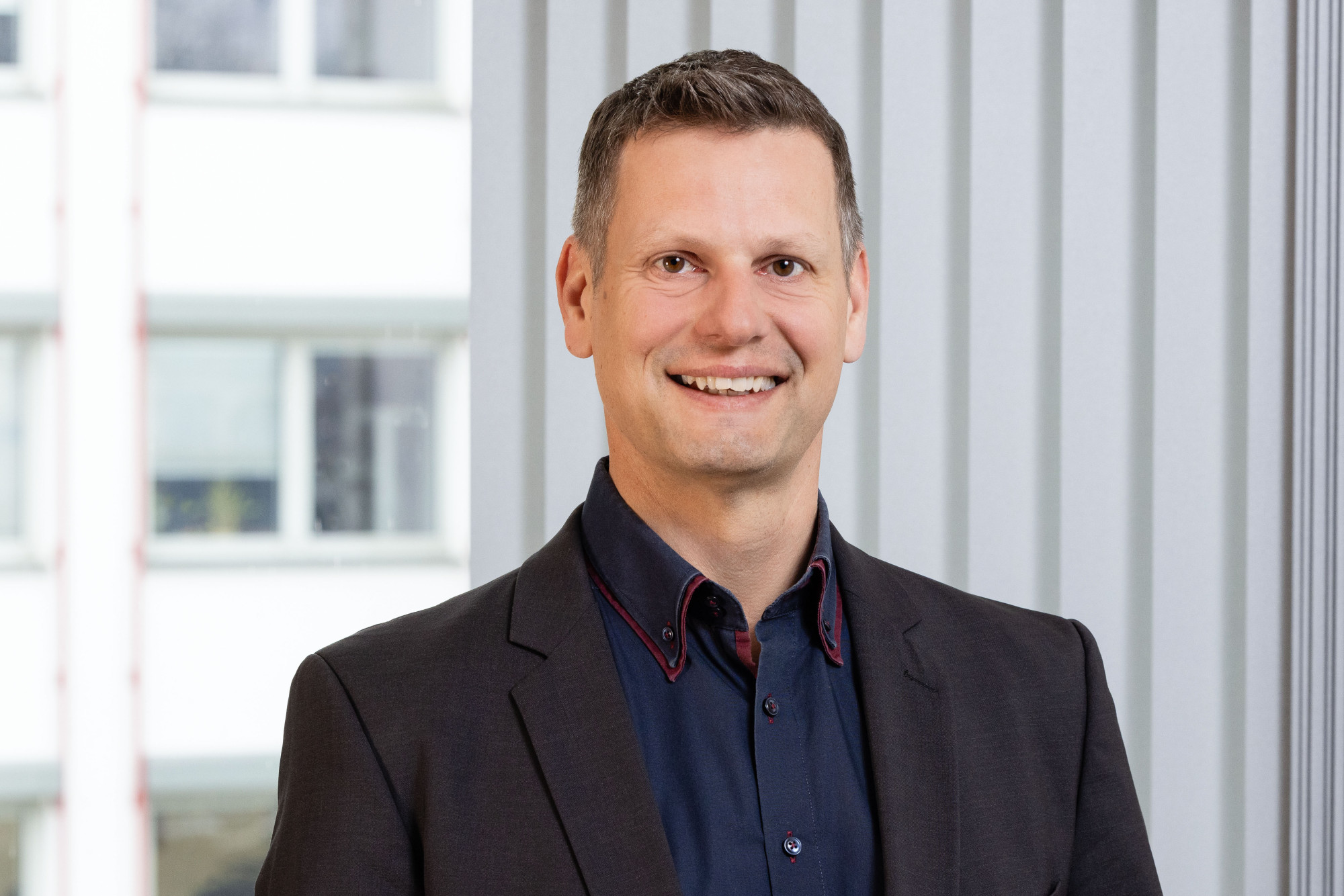 Ein lächelnder Mann in einem dunklen Blazer und einem hellen Hemd steht vor einem modernen Hintergrund mit vertikalen Linien.